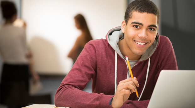 A student sits behind his laptop and holds a pencil.